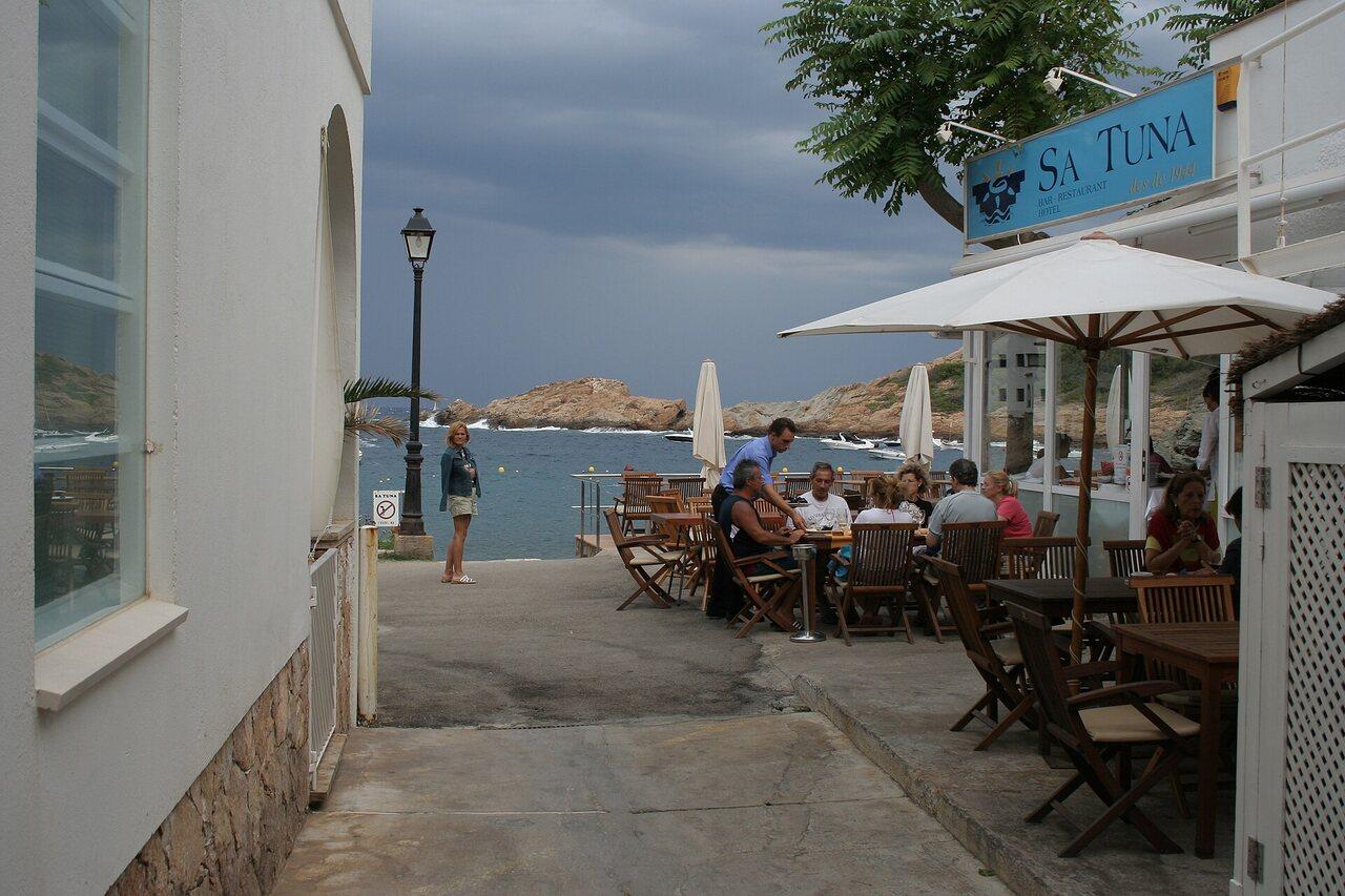 Narrow waterfront lane with restaurant tables beside Sa Tuna cove